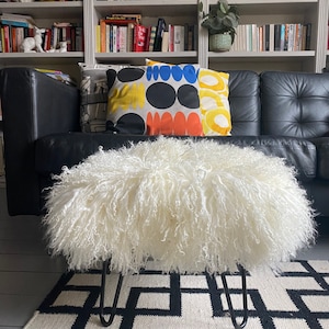 May include: A white fluffy sheepskin ottoman with black hairpin legs sits on a black and white geometric rug in front of a black leather sofa with colorful throw pillows.