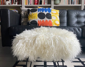 May include: A white fluffy sheepskin ottoman with black hairpin legs sits on a black and white geometric rug in front of a black leather sofa with colorful throw pillows.