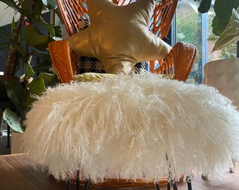 May include: A wicker chair with a fluffy white sheepskin footstool and a gold star-shaped pillow. The chair is in a room with plants and a patterned wall.