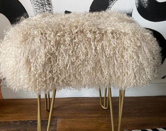 May include: A cream-colored, fluffy ottoman with a rounded top and gold hairpin legs. The ottoman has a textured, curly surface. The legs are thin and angled, providing a modern touch to the design. The background features a black and white abstract pattern.