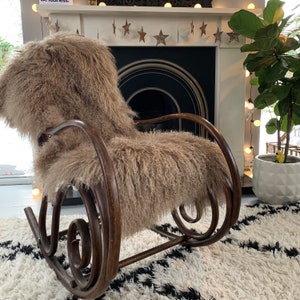May include: A wooden rocking chair with a brown faux fur cushion. The chair is in front of a fireplace with a white mantel and a string of lights.