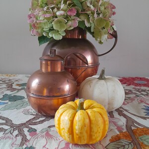 May include: A copper jug and a round copper container with a lid are arranged with two small pumpkins and a bouquet of pink and green flowers. The pumpkins are white and yellow. The objects are on a floral patterned tablecloth.