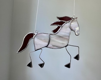 May include: A stained glass suncatcher shaped like a horse in motion. The horse features a white, opalescent body with dark red glass accents on the mane and tail. The pieces are joined by dark metal solder lines, and it hangs from thin white strings.