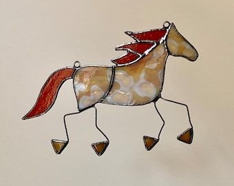 Pode incluir: Um apanhador de sol de vitral em forma de cavalo em movimento. O corpo apresenta vidro marmoreado creme e branco, enquanto a crina e a cauda são de vidro texturizado vermelho-alaranjado. As pernas são fios metálicos finos com cascos triangulares.