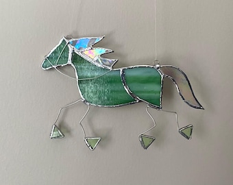 Pode incluir: Um ornamento de cavalo em vitral com painéis de vidro texturizado verde no corpo e na cabeça, e detalhes em vidro iridescente na crina e na cauda. A peça é construída com linhas de solda prateadas e pernas de arame fino que terminam em pequenos cascos de vidro triangulares.