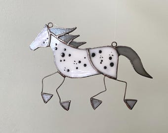 Pode incluir: Um ornamento de cavalo em vitral com vidro branco e manchas pretas, crina e cauda cinzentas, e pernas de arame metálico fino com cascos triangulares. A peça inclui dois pequenos anéis de metal na parte superior para a pendurar facilmente.