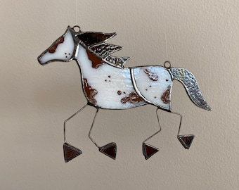 Pode incluir: Um ornamento de vitral em forma de cavalo com painéis de vidro manchado em branco e castanho, unidos por linhas de soldadura de metal escuro. O cavalo tem uma crina castanho-escura, uma cauda cinzenta texturizada e quatro pernas de metal finas.