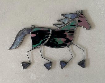 Pode incluir: Um ornamento de cavalo em vitral com um corpo de painéis de vidro iridescente preto, verde e rosa. A crina e a cauda são feitas de vidro translúcido roxo e azul. As pernas são fios de metal finos que terminam em pequenos pés de vidro triangulares.