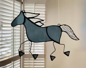 May include: A stained glass horse suncatcher featuring a dark blue body with a textured clear glass mane and tail. The horse is constructed with black metal solder lines and includes thin wire legs with triangular feet, suspended by white strings.