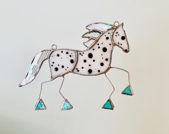 May include: A stained glass horse ornament featuring a white body with black spots, a shimmering iridescent mane and tail, and four legs ending in small triangular turquoise glass hooves, all held together by thin silver metal solder lines.
