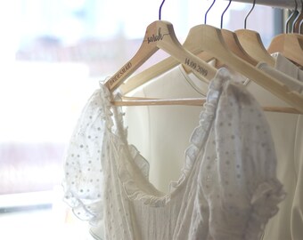 May include: A white eyelet dress hangs on a wooden hanger labeled "Bridesmaid" and "Sarah". The hanger also has the date "14.09.2019" printed on it. Other white garments are visible on hangers in the background.