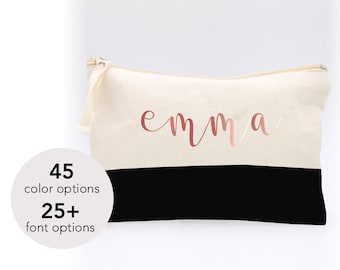 May include: A canvas cosmetic bag with a two-tone design. The top half is a cream color, and the bottom half is black. The name "emma" is written in a rose gold script across the cream section. The bag has a zipper closure and a small loop on the side.