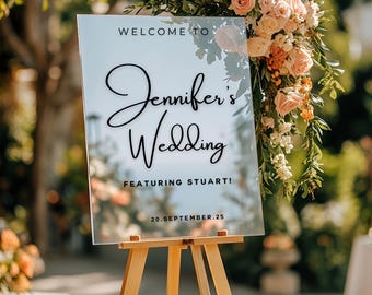 May include: A clear acrylic wedding sign on a wooden easel. The sign reads "WELCOME TO Jennifer's Wedding FEATURING STUART! 20.SEPTEMBER.25" in elegant black script. The background features a floral arrangement with pink and orange roses.