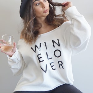 May include: Off-the-shoulder white sweatshirt with the words "WINELOVER" printed in black. The person is holding a wine glass with a pink drink and the words "thank you" in gold. The person is wearing a black hat and black pants.