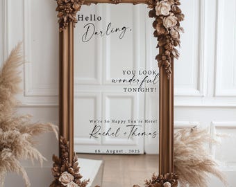May include: A large, ornate, rectangular mirror with a brown, floral-carved frame. The mirror has the text "Hello Darling, You look wonderful tonight! We're So Happy You're Here! Rachel + Thomas 06 August 2025". The frame is decorated with flowers and foliage.