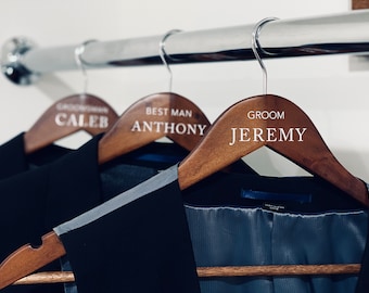 May include: Three wooden suit hangers with silver hooks, each holding a dark suit. The hangers are labeled with white text: "Groom Jeremy", "Best Man Anthony", and "Groom Caleb". The suits hang on a chrome rod.