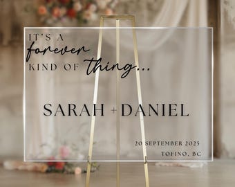 May include: A clear acrylic wedding sign on a gold easel. The sign reads "IT'S A forever KIND OF thing... SARAH + DANIEL 20 SEPTEMBER 2025 TOFINO, BC" in black script and block lettering. The background is blurred with floral arrangements.