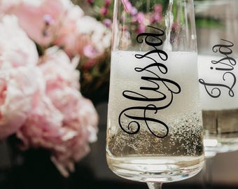 May include: Two champagne flutes, one filled with bubbly and the other partially filled. The word "Alyssa" is written in elegant script on the front of the first glass, and "Lisa" on the second. Pink flowers are in the background.