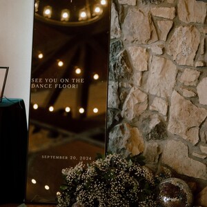 May include: A tall, rectangular mirror with the initials "E A" and the text "SEE YOU ON THE DANCE FLOOR! SEPTEMBER 20, 2024" is propped against a stone wall. The mirror reflects lights and is decorated with flowers and disco balls.