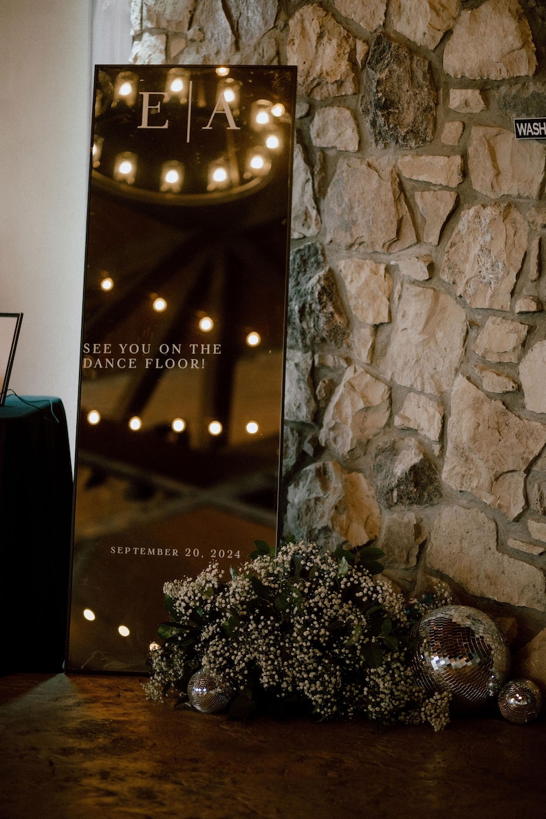 May include: A tall, rectangular mirror with the initials "E A" and the text "SEE YOU ON THE DANCE FLOOR! SEPTEMBER 20, 2024" is propped against a stone wall. The mirror reflects lights and is decorated with flowers and disco balls.