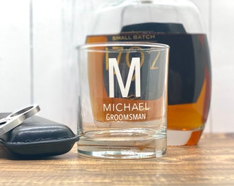 May include: A personalized rocks glass with the letter "M" and the name "MICHAEL GROOMSMAN" in white lettering. A bottle of liquor is in the background. A black leather case and a metal bottle opener are in the foreground.