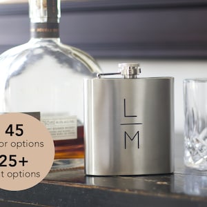 May include: A silver-coloured hip flask with the initials "LM" in black. A clear glass and a bottle of liquor are in the background. The image also includes text that says "45 colour options" and "25+ font options".