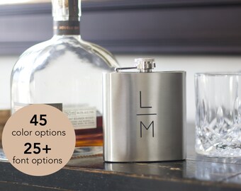 May include: A stainless steel flask with the initials "LM" in black. A clear glass and a bottle of liquor are in the background. The image also includes text that says "45 color options" and "25+ font options".