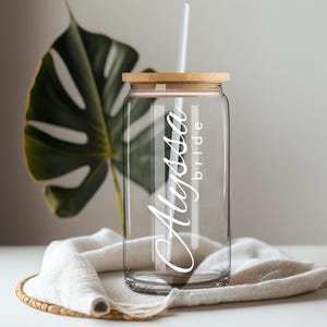 May include: A clear glass tumbler with a bamboo lid and a white straw. The glass features the white text Alyssa in a large script font and the word bride in a smaller sans-serif font printed vertically along the side of the cylindrical vessel.
