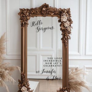 May include: A large, ornate, rectangular mirror with a brown frame. The frame is decorated with floral and leaf details. The mirror has the text "Hello Gorgeous" and "You look incredible. Now let's celebrate!" written on it.