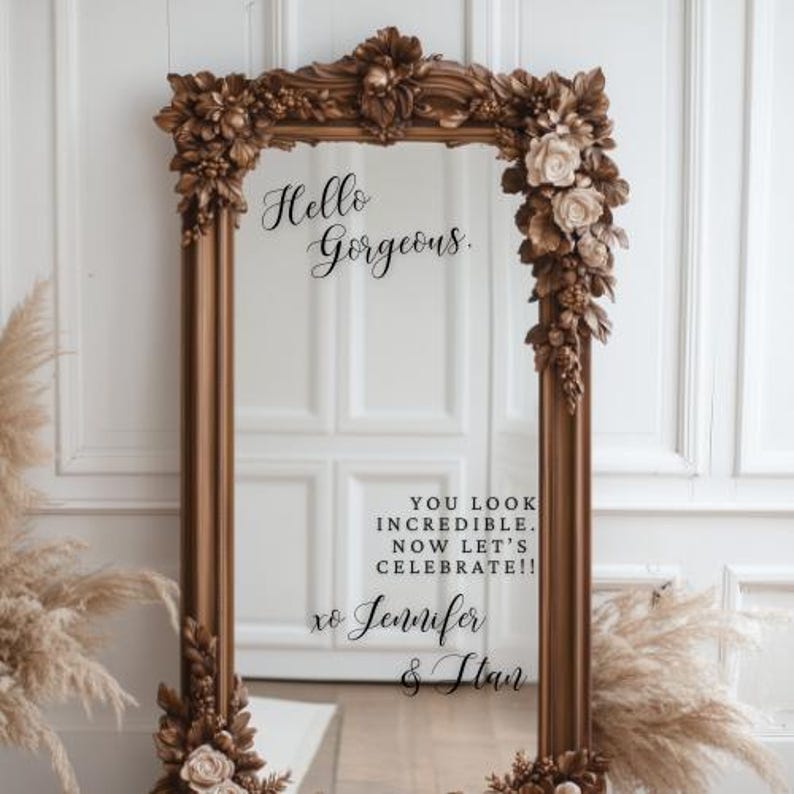 May include: A large, ornate, rectangular mirror with a brown frame. The frame is decorated with floral and leaf details. The mirror has the text "Hello Gorgeous" and "You look incredible. Now let's celebrate!" written on it.
