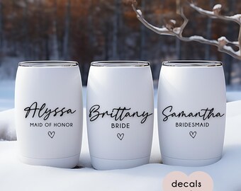 May include: Three white stemless wine tumblers with black text. The first reads "Alyssa, Maid of Honor" with a heart. The second reads "Brittany, Bride" with a heart. The third reads "Samantha, Bridesmaid" with a heart. The background is a snowy outdoor scene.