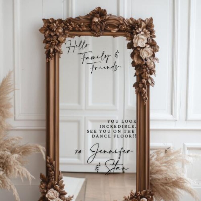 May include: A large, ornate, rectangular mirror with a brown, floral-decorated frame. The mirror's surface reflects a white door. Text on the mirror reads "Hello Family & Friends," "You look incredible. See you on the dance floor!!" and "to Jennifer & Stan."
