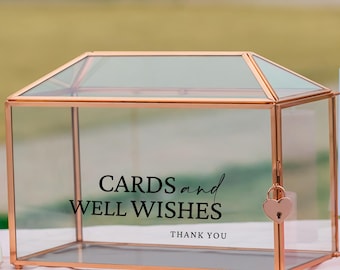May include: A clear glass card box with a copper-colored metal frame and a heart-shaped lock. The box is inscribed with the words "CARDS and WELL WISHES THANK YOU" in black script, ideal for weddings or special events.