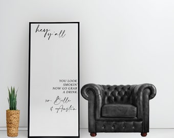 May include: A black framed sign with the text "hey y'all" at the top and "YOU LOOK SMOKIN' NOW GO GRAB A DRINK. xo. Belle & Austin" below. A black leather tufted armchair is to the right. A potted plant is on the left.