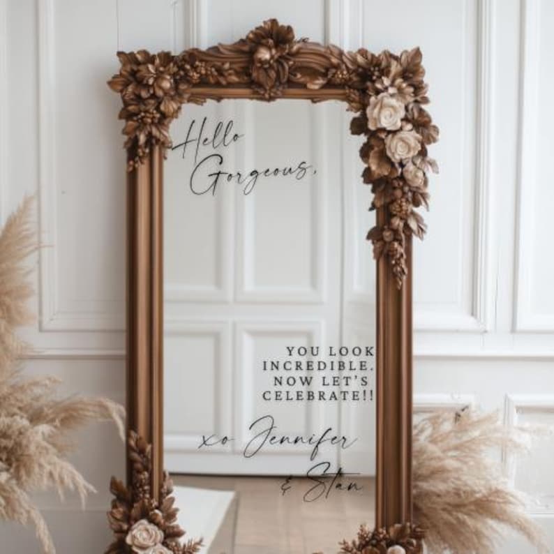 May include: A large, ornate, rectangular mirror with a brown frame. The frame is decorated with floral and leaf accents. The mirror has the text "Hello Gorgeous" and "You look incredible. Now let's celebrate!!" written on it.