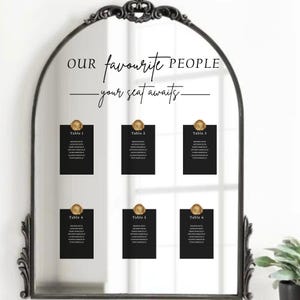 May include: A black framed mirror with the text "Our favourite PEOPLE - your seat awaits". Six black cards with gold wax seals are labeled "Table 1" through "Table 6", each listing names. A decorative wedding or event seating chart.