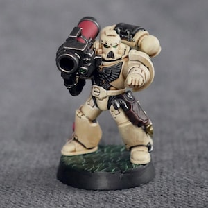 Puede incluir: Una miniatura de plástico beige y negro de un marine espacial sosteniendo un lanzacohetes. La miniatura está pintada con detalles en negro, rojo y verde.