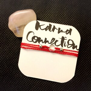 Puede incluir: Pulsera de cuerda roja con un dije hexagonal plateado y cuentas plateadas. La pulsera está sobre una tarjeta blanca con el texto "Karma Connection" escrito en negro.