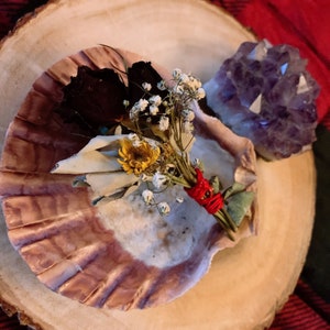May include: A seashell bowl holds a dried flower bundle with a red string, a dried rose, and a yellow flower. A purple amethyst crystal sits beside the bowl on a wooden surface. The background is a red and black plaid.
