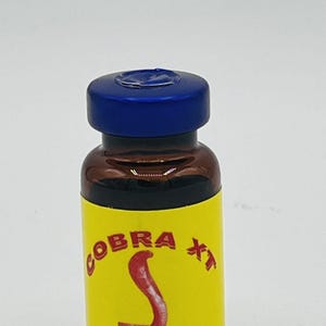 May include: A small brown glass bottle with a blue cap. The bottle has a yellow label with red text that says "COBRA XT" and a red image of a cobra. The label also says "FORMULACI&Oacute;N COMPLETA".