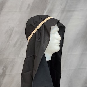 May include: A black head covering featuring a long, draped fabric design that extends down the back and sides. The headpiece is secured with a light beige, elasticized band that encircles the crown, creating a structured fit around the head.