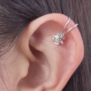 Puede incluir: Un pendiente de oreja de plata con un pequeño diseño de tortuga. El pendiente está hecho de alambre y está diseñado para envolverse alrededor de la parte superior de la oreja.