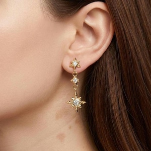 May include: Gold-toned dangle earrings featuring a star and diamond design. The earrings have a star-shaped stud, a small diamond, and a larger starburst pendant with a central diamond. The earrings are worn on the ear.