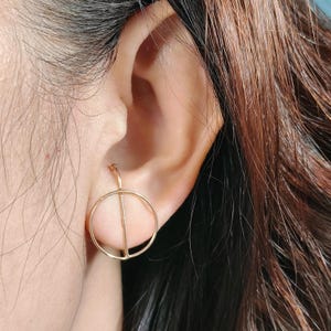 Puede incluir: Pendientes de aro de oro con una línea fina en el centro. Los pendientes se llevan en la oreja de una persona.