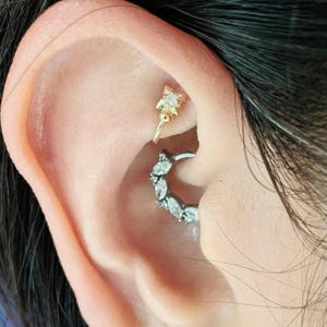 Pode incluir: Um close-up de uma orelha de uma pessoa com dois piercings. O piercing inferior é um aro com pequenas pedras transparentes. O piercing superior é uma estrela dourada com uma pedra transparente no centro.