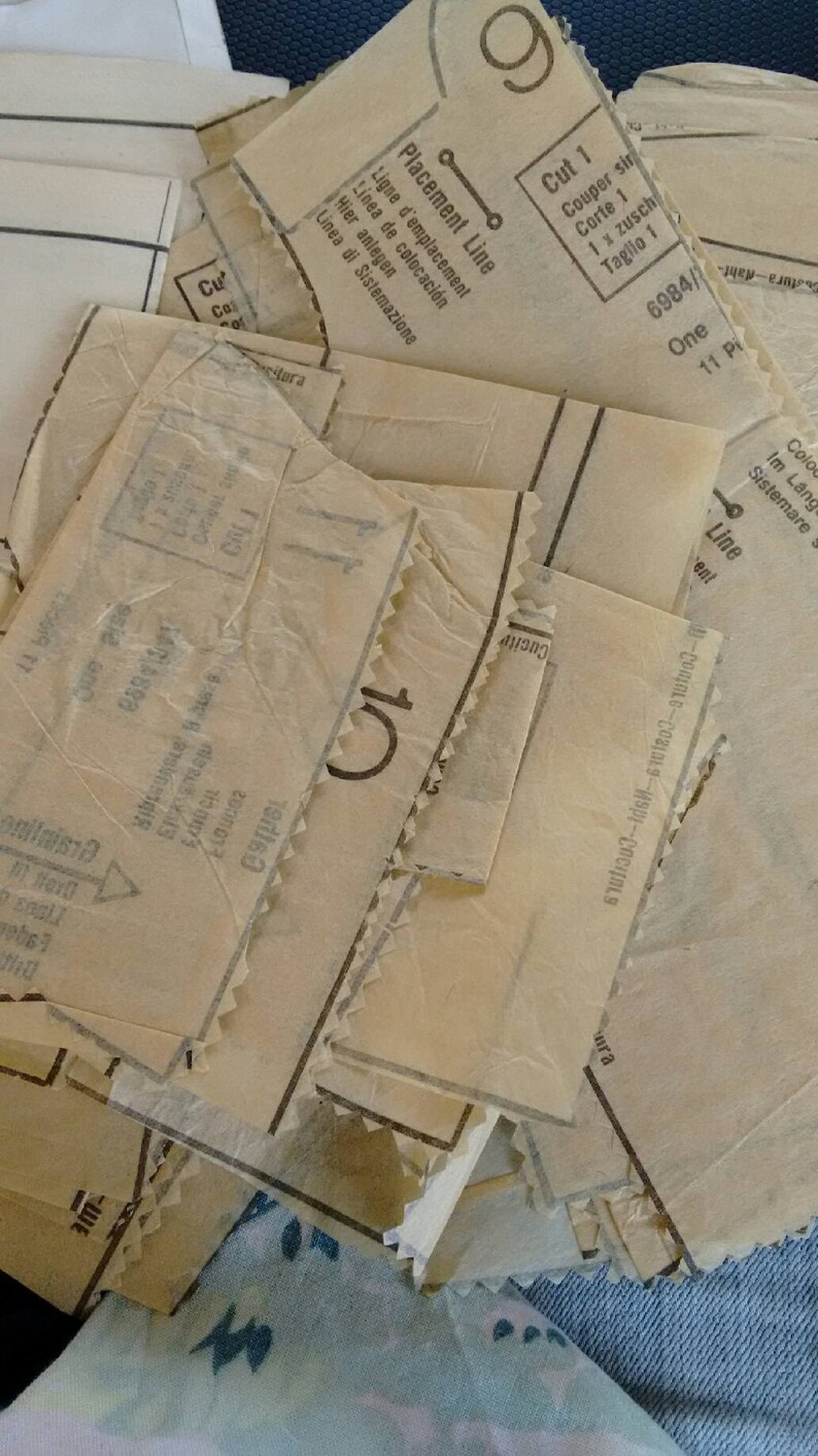 Pu&ograve; includere: Primo piano di una pila di pezzi di cartamodello vintage su uno sfondo di denim blu. I pezzi del cartamodello sono etichettati con istruzioni in pi&ugrave; lingue, tra cui inglese, francese, tedesco, italiano e spagnolo. I pezzi del cartamodello sono per un capo con una linea di posizionamento e un'istruzione di taglio 1.