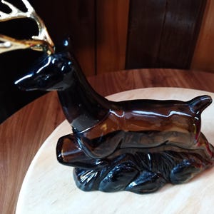 May include: A dark brown glass deer figurine with gold antlers. The deer is standing on a black base and is in a crouching position.