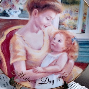 Könnte beinhalten: Dekorative Platte mit Goldrand, die eine Mutter und ein Kind zeigt, mit dem Text "Mother's Day 1995". Die Platte wird auf einem dekorativen Ständer präsentiert. Das Bild zeigt eine Frau in einem gelben Kleid, die ein Kind hält.