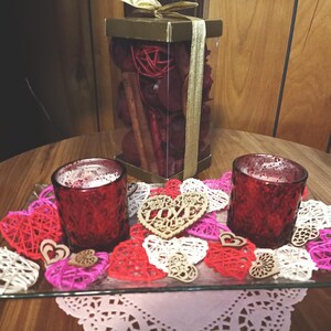 May include: Two red glass candle holders with white wax inside, surrounded by pink, white and brown heart-shaped decorations on a glass tray. The tray is on a wooden table with a box of potpourri in the background.