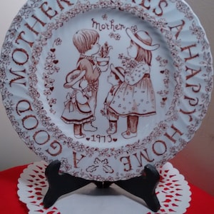 May include: A decorative plate with a brown and white design. The plate features an illustration of children and the text "GOOD MOTHER MAKES A HAPPY HOME" and "1973". The plate is displayed on a black stand.